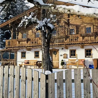 Ein gemütliches Holzhaus im Schnee, umgeben von einem Holzlattenzaun. Die Szenerie vermittelt eine ruhige Winteratmosphäre.