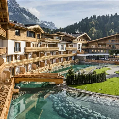 Ein schönes Wellness-Resort mit Holzarchitektur und einem klaren Wasserlauf. Im Hintergrund sind die Berge und ein grüner Bereich mit Rasen und Pflanzen zu sehen.