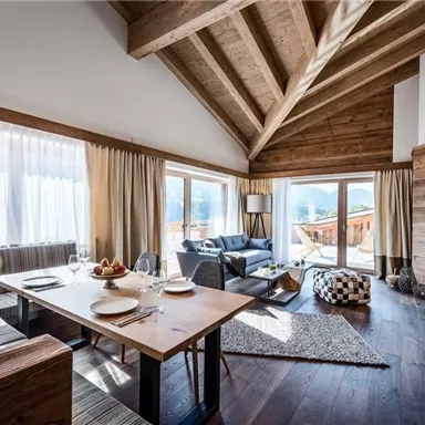 A modern living room with a wooden ceiling and large windows. The space is furnished with a dining table, comfortable seating, and cozy decor.