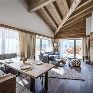 Ein modernes Wohnzimmer mit Holzelementen und großen Fenstern. Es gibt einen Esstisch, eine gemütliche Sitzecke und viel Tageslicht.