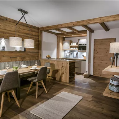 Ein gemütlicher Wohnraum im modernen Landhausstil mit Holzdetails. Es gibt einen Essbereich mit einem großen Tisch und modernen Stühlen sowie eine offene Küche.