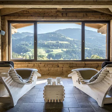 Eine gemütliche Lounge mit zwei Liegestühlen und einem atemberaubenden Ausblick auf die Berge. Große Fenster lassen viel Tageslicht herein und schaffen eine einladende Atmosphäre.