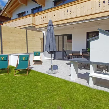 Eine gemütliche Terrasse mit einem Grill und Liegestühlen. Umgeben von einem gepflegten Rasen und einer modernen Holzverkleidung.