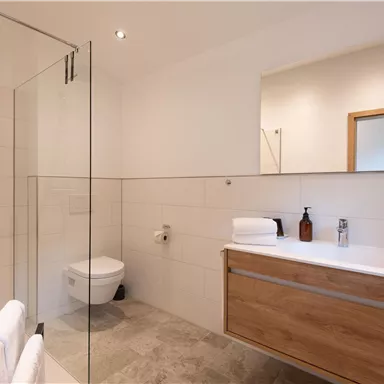 Ein modernes Badezimmer mit einer Glasdusche und einem Holzschrank. Es sind frische Handtücher und Pflegeprodukte sichtbar.