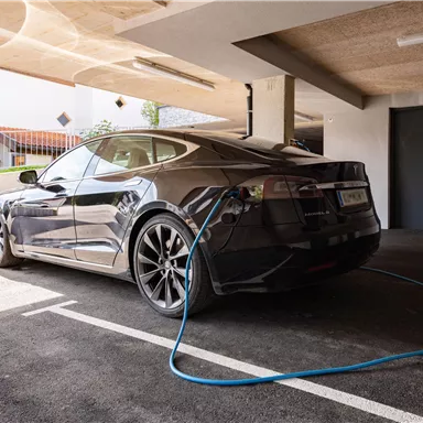 Ein schwarzes Elektroauto steht in einer Garage und ist an eine Ladestation angeschlossen. Im Hintergrund sind Fenster und eine Tür sichtbar.
