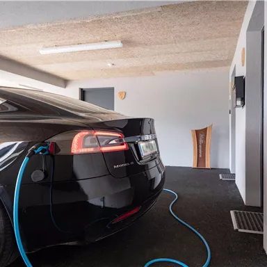Ein schwarzes Elektroauto wird in einer modernen Garage aufgeladen. Im Hintergrund sind neutrale Wände und einige Möbelstücke zu sehen.