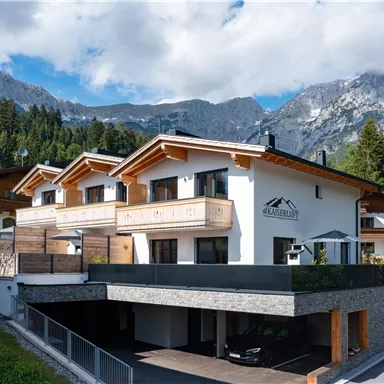 Ein modernes Haus in den Bergen mit Holzverkleidung und Balkonen. Im Hintergrund sind beeindruckende Berge und ein blauer Himmel zu sehen.