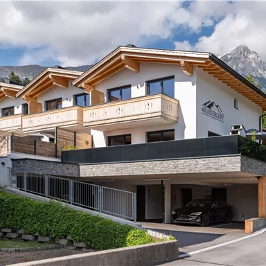 Ein modernes Gebäude mit Holzbalustraden und einem Garagenbereich. Im Hintergrund sind Berge und eine grüne Umgebung zu sehen.