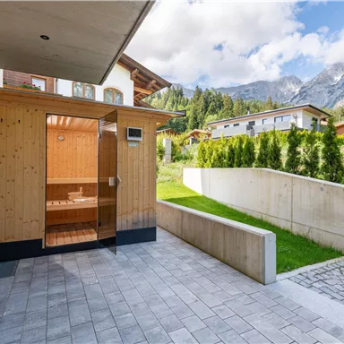 Ein moderner Saunabereich aus Holz in einem schönen Garten. Im Hintergrund sind Berge und ein klarer Himmel sichtbar.