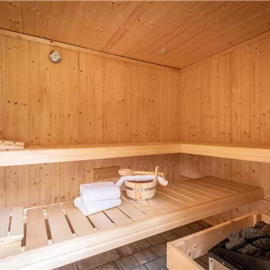 Eine gemütliche Sauna aus Holz mit Bänken und einem kleinen Ofen. In der Ecke liegen frische Handtücher und ein Holzbehälter.