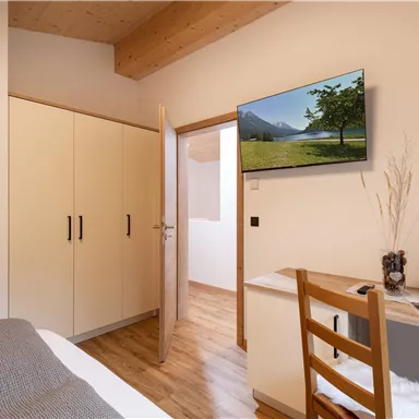 Ein gemütliches Zimmer mit einem Bett, einem Schreibtisch und einem Fernseher an der Wand. Die Einrichtung ist modern und hell, mit einem Holzfußboden und einem Schrank.