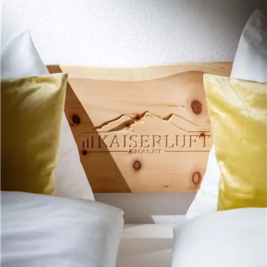 Ein komfortables Bett mit einer hölzernen Kopfstütze und dem Logo "KAISERLUFT". Es gibt dekorative Kissen in Gelb und weiße Bettwäsche.