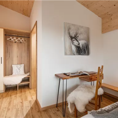 Ein helles Zimmer mit Holzboden und moderner Einrichtung. Auf einem kleinen Tisch steht ein Buch, daneben ein Stuhl mit einem gemütlichen Fell.