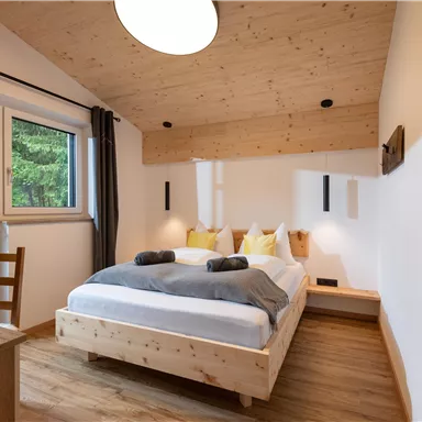 Ein modernes Schlafzimmer mit einem Doppelbett und Holzdetails. Große Fenster sorgen für viel Tageslicht und einen schönen Blick ins Grüne.