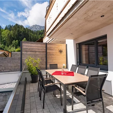 Eine moderne Terrasse mit einem großen Tisch und Stühlen. Umgeben von grünen Bäumen und einem blauen Himmel.