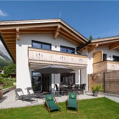 Eine moderne Berghütte mit großzügiger Terrasse und Garten. Im Hintergrund sind majestätische Berge und blauer Himmel zu sehen.
