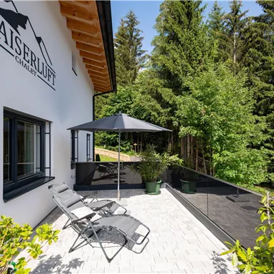 Eine moderne Terrasse mit Liegen und einem Sonnenschirm. Umgeben von üppigem Grün und Bäumen wirkt der Ort einladend und ruhig.