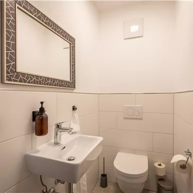 Ein modernes Badezimmer mit einem Waschbecken und einem WC. An der Wand befindet sich ein Spiegel und die Fliesen sind hell und schlicht.