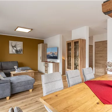 Ein modernes Wohnzimmer mit einem bequemen grauen Sofa und einem holzbedeckten Tisch. Der Raum hat eine warme Atmosphäre und eine klare, helle Einrichtung.
