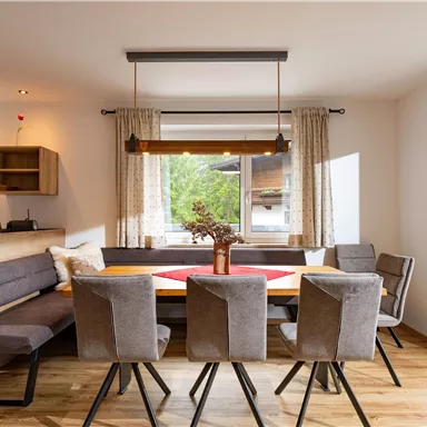 Ein modernes Esszimmer mit einem großen Tisch und grauen Stühlen. Große Fenster lassen viel Tageslicht herein und bieten einen Blick ins Grüne.