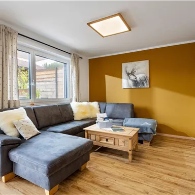 Ein modernes Wohnzimmer mit einem grauen Sofa und einem Holztisch. Die Wände sind teilweise orange gestrichen, und es gibt große Fenster mit Blick nach draußen.