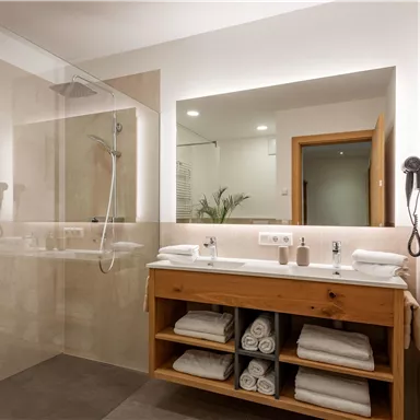 A modern bathroom with a glass shower cubicle and a wooden sink. Fresh towels and toiletries are neatly arranged on the shelf.