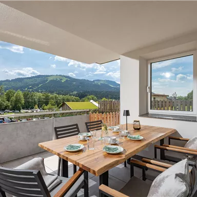 A cozy terrace with a wooden table and chairs. In the background, green mountains and a clear sky can be seen.