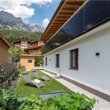 A quiet garden with loungers and a modern, white wall. In the background, mountains and other buildings are visible.