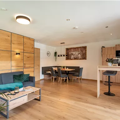 A modern furnished living area with wood paneling and an open kitchen. There is a cozy seating area and a dining table for four people.