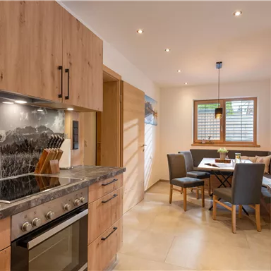 A modern kitchen with wooden cabinets and a cooking area. In the background, there is a dining area with a table and chairs.