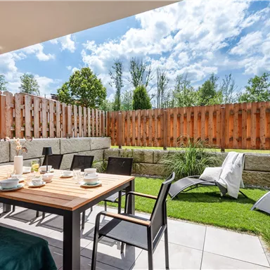 A cozy terrace with a table, chairs, and a beautiful view of the garden. The sun is shining and the sky is partly cloudy.