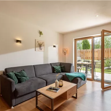 A modern living room with a gray sofa and a wooden table. Large windows offer a view of the garden.