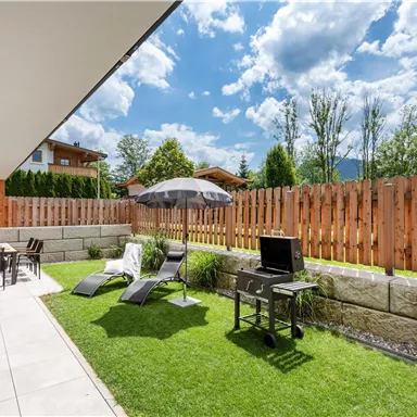 A cozy garden with grass, a sun umbrella, and loungers. In the background, a wooden terrace and a grill can be seen.