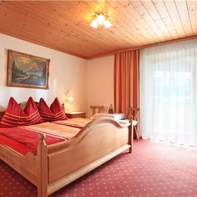 Ein gemütliches Schlafzimmer mit einem Doppelbett und roten Kissen. Große Fenster sorgen für viel Tageslicht.