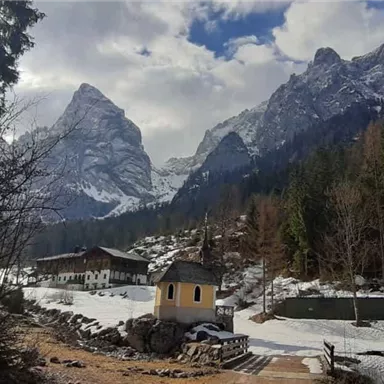 Eine malerische Berglandschaft mit schneebedeckten Gipfeln. Im Vordergrund steht ein kleines Haus und es gibt grünes Umland.