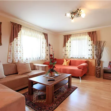 Ein helles Wohnzimmer mit einem beigen Sofa und einem roten Sofa. Große Fenster lassen viel Licht herein, und ein Tisch steht in der Mitte des Raumes.