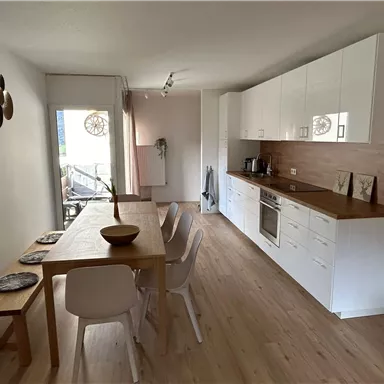 A modern kitchen with a wooden dining area. Bright walls and large windows create an inviting atmosphere.