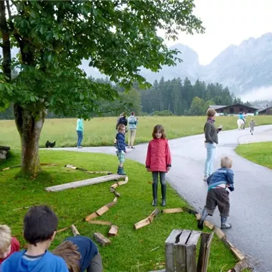 A group of children is playing outdoors, surrounded by trees and mountains. Some children are building with wooden pieces while others are active nearby.