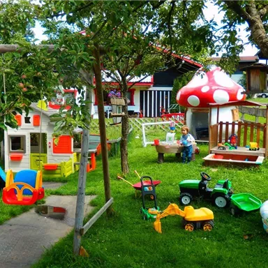 Ein bunter Spielplatz im Garten mit verschiedenen Spielzeugen und einem Spielhaus. Im Hintergrund sitzt ein Kind und spielt unter einem Baum.