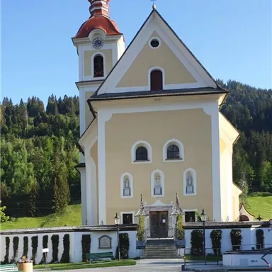 A picturesque church with a red tower and yellow facade. Surrounded by green hills and trees.
