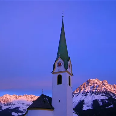 A church with a tall, pointed tower stands under a colorful sky. Snow-covered mountains can be seen in the background.