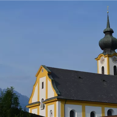 Eine hübsche Kirche mit einer gelben Fassade und einer markanten Spitze. Im Hintergrund sind Berge und ein blauer Himmel zu sehen.