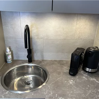 A modern kitchen corner with a round stainless steel sink and a black faucet. Next to the sink are a coffee maker and a kettle.