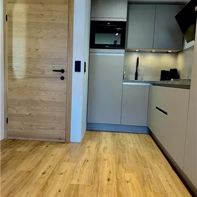 A modern kitchen with bright furniture and wooden flooring. The wall is partially equipped with gray cabinets and a cooking surface.