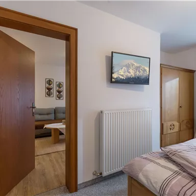Ein gemütliches Schlafzimmer mit einem Bett und einem Fernseher an der Wand. Durch die offene Tür ist ein Wohnbereich zu sehen.