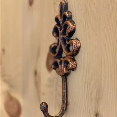 Ein dekorativer Metallhaken an einer Holzwand. Der Haken hat ein kunstvolles Design und ist in Kupferfarbe gehalten.