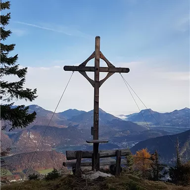 Ein Holzkreuz auf einem Gipfel mit weitem Blick auf die Berge. Der Himmel ist blau, und die Landschaft zeigt sanfte Hügel und Wälder.