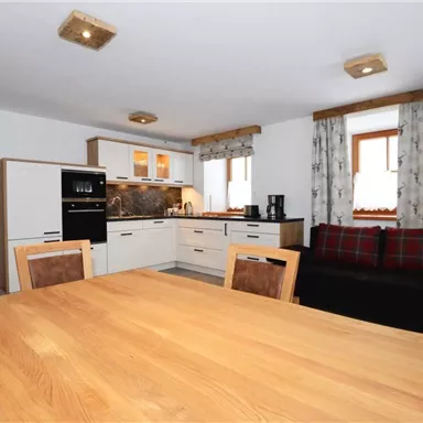 A modern kitchen with a large wooden dining table. The room is bright and cheerfully designed, featuring a cozy sofa and windows with curtains.