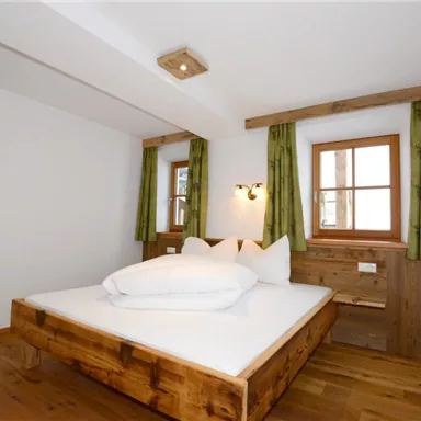 A modern bedroom with a large wooden bed and white pillows. The walls are bright, and the windows have green curtains.
