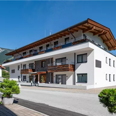 A modern building with several apartments, surrounded by trees and a clear blue sky. In the background, gentle mountains can be seen.